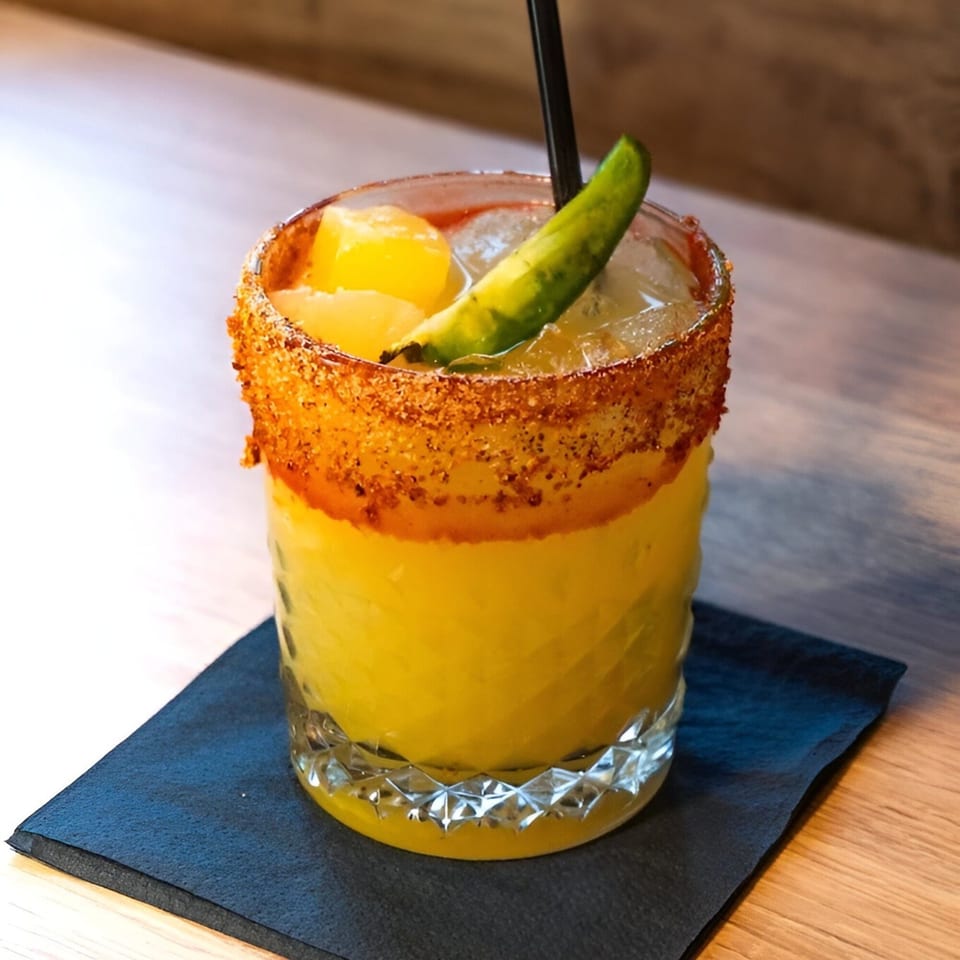A vibrant yellow cocktail with a chili-salt rim, garnished with a green pepper and pineapple chunks.