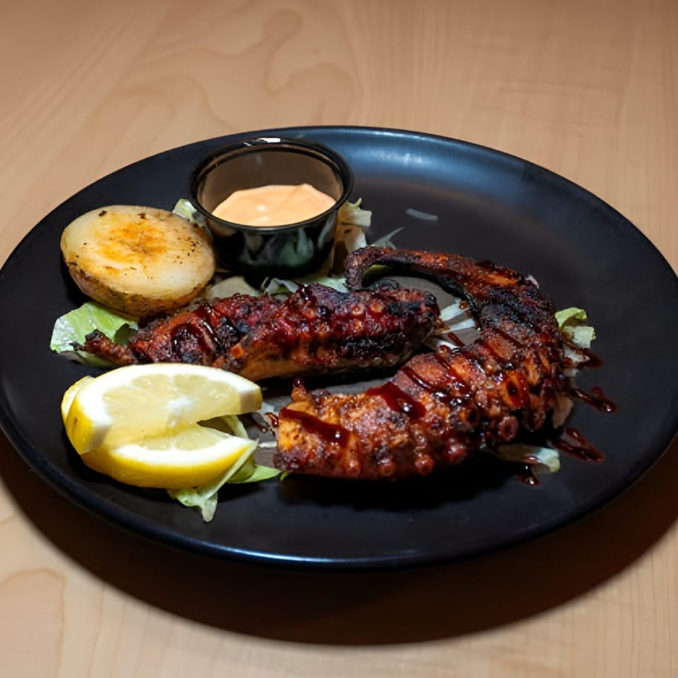 Grilled octopus with a side of roasted potato, lemon wedges, and a dipping sauce on a black plate.